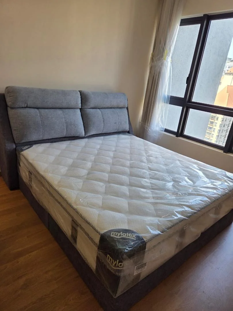 Bedroom features: queen-size bed, upholstered headboard, mattress with protective cover, wooden flooring, floor-to-ceiling window, sheer curtains, grey fabric bed frame, decorative wall, natural light from window; style: modern; view: city.